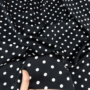 Puede incluir: Tela negra con lunares blancos. La tela está drapeada, mostrando la textura y el patrón. Los lunares blancos están espaciados uniformemente sobre el fondo negro, creando un diseño clásico.