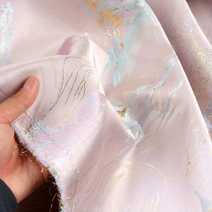 Könnte beinhalten: Nahaufnahme eines blassrosa Stoffes mit schimmernder, strukturierter Oberfläche. Der Stoff weist ein zartes Muster aus goldenen, hellblauen und lavendelfarbenen Akzenten auf. Das Material ist weich und fließend, mit einem dezenten Glanz.