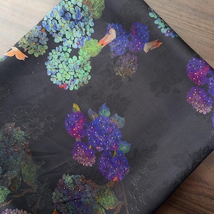 May include: Dark fabric with a floral design. The pattern features clusters of blue, purple, and green hydrangea flowers. The fabric has a subtle sheen and a dark background, with detailed floral and leaf designs.