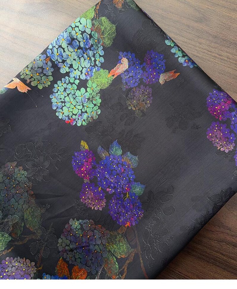 May include: Dark fabric with a floral design. The pattern features clusters of blue, purple, and green hydrangea flowers. The fabric has a subtle sheen and a dark background, with detailed floral and leaf designs.