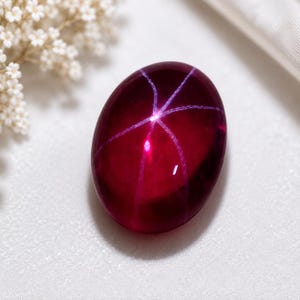 Könnte beinhalten: Ein tiefroter, ovaler Edelstein mit einem markanten sechsstrahligen Sterneffekt, bekannt als Asterismus. Der im Cabochon-Schliff gearbeitete Stein zeigt eine glatte, polierte Oberfläche mit einem Lichtreflex, der ein kreuzartiges Muster bildet.