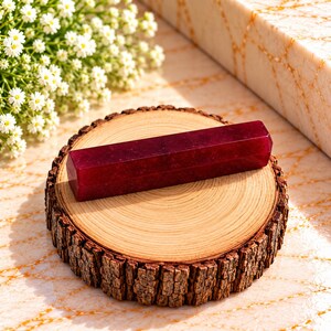 May include: A deep red, hexagonal crystal wand resting horizontally on a circular wooden slice with visible tree rings and rough bark edges. The crystal has a smooth, polished surface with subtle internal inclusions and a dark, saturated colour.