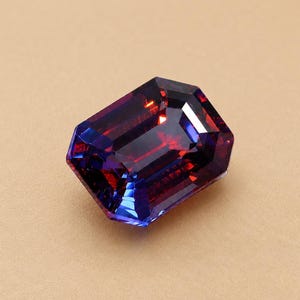 Puede incluir: Una piedra preciosa rectangular de corte esmeralda que presenta un vibrante juego de colores cambiantes en tonos rojo intenso, morado y azul. La superficie facetada refleja la luz con gran claridad, mostrando bordes definidos.