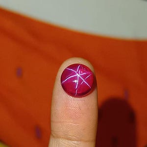 May include: A round, cabochon-cut star ruby gemstone featuring a deep magenta-red color and a distinct, bright white six-rayed star pattern that radiates from the center of the stone's smooth, polished, and domed surface.