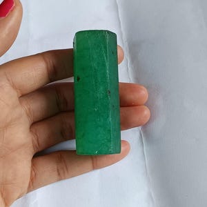 Puede incluir: Una piedra cilíndrica de color verde esmeralda opaco con una superficie rugosa y texturizada e inclusiones oscuras visibles. La piedra presenta una forma hexagonal con extremos planos, mostrando un color verde profundo y saturado.