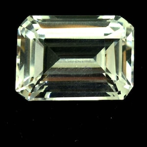 Pode incluir: Uma pedra preciosa de corte retangular com um tom amarelo claro. A gema tem uma superfície facetada que reflete a luz, criando um efeito brilhante. A pedra preciosa está sobre um fundo preto, destacando sua clareza. Este é um componente de joalheria.