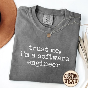 May include: A charcoal grey short-sleeved t-shirt with a crew neckline and the white printed text "trust me, i'm a software engineer" on the front. The shirt features a Comfort Colors brand label on the inside collar indicating a size small.