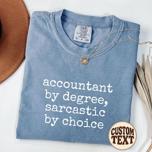 Può includere: Una t-shirt blu a maniche corte con scollo rotondo e una stampa grafica bianca che recita: accountant by degree, sarcastic by choice. La maglietta presenta un'etichetta del marchio Comfort Colors sul colletto interno e una collana dorata.