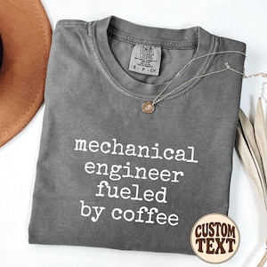 May include: A charcoal gray crew neck t-shirt featuring the white printed text "mechanical engineer fueled by coffee" on the front. The shirt has a Comfort Colors brand label at the inner neckline indicating size small and is made of 100% cotton.