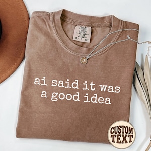 May include: A brown short-sleeved t-shirt with a crew neckline featuring the white text "ai said it was a good idea" printed on the front. The shirt has a Comfort Colors brand label at the inner neck showing size S-CH and a small circular logo.