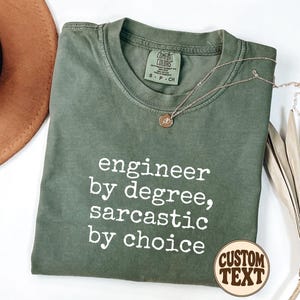 May include: An olive green crew neck t-shirt with white text that reads "engineer by degree, sarcastic by choice." The shirt features a Comfort Colors brand label at the inner neckline and is displayed with a gold necklace and a hat.