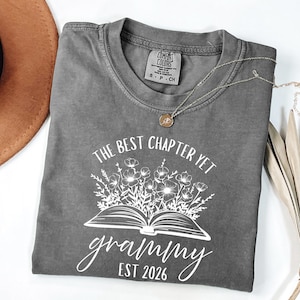 Może przedstawiać: Szara koszulka Comfort Colors z białym napisem "THE BEST CHAPTER YET grammy EST 2026". Projekt zawiera otwartą książkę z kwiatowymi akcentami. Widoczny jest również złoty naszyjnik i brązowy kapelusz.