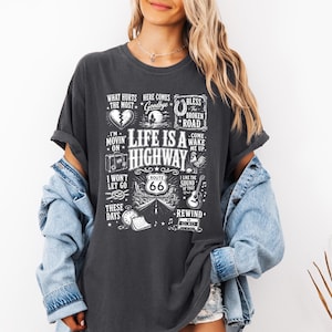 Puede incluir: Una camiseta de manga corta de color gris carbón con un estampado gráfico blanco con el texto "LIFE IS A HIGHWAY" y "ROUTE 66" rodeado de varias ilustraciones que incluyen un corazón roto, una guitarra, un casete y un reloj.