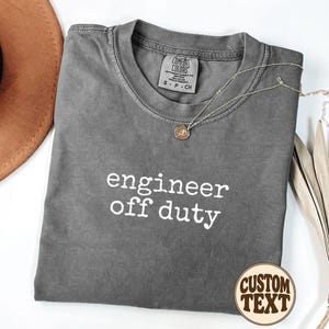 May include: A charcoal grey short-sleeve t-shirt with a crew neckline featuring the white text "engineer off duty" printed on the chest. The shirt has a Comfort Colors brand label at the inner neck and is displayed with a gold pendant necklace.