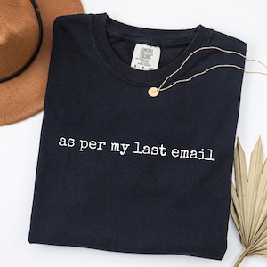 May include: A navy blue Comfort Colors crew neck t-shirt with the phrase "as per my last email" in white. A gold necklace and a brown hat are also in the image. A dried palm leaf is in the bottom right corner.