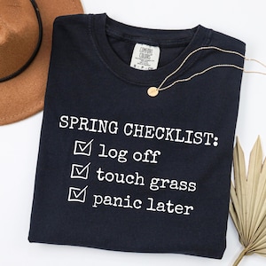 May include: A navy blue t-shirt with the text "SPRING CHECKLIST: log off, touch grass, panic later" in white. A gold necklace and a brown hat are also in the image. The shirt is made of a soft material.