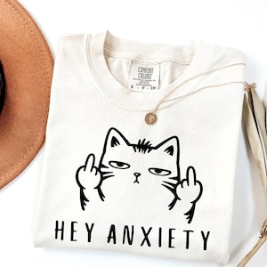 May include: Cream-colored t-shirt with a black graphic of a cat giving the middle finger. The text "HEY ANXIETY" is below the cat. A gold necklace and a brown hat are visible in the background.