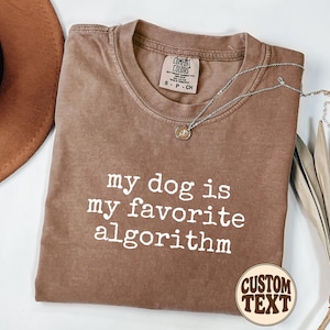 May include: A brown short-sleeve t-shirt with the white text "my dog is my favorite algorithm" printed on the front. The shirt features a crew neckline and a Comfort Colors brand label on the inside collar. A silver necklace rests on the shirt.