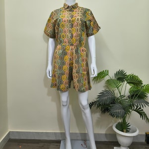 May include: A two-piece outfit on a mannequin. The set includes a short-sleeved collared shirt and matching shorts. The fabric features a repeating pattern of colorful floral and geometric shapes in shades of yellow, pink, green, and brown. The outfit is displayed on a white mannequin.