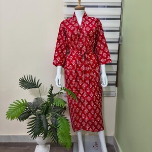 May include: A red patterned robe with white floral designs and white trim is displayed on a mannequin. The robe has long sleeves and a matching belt. The mannequin is positioned in front of a window with blinds and a potted plant.