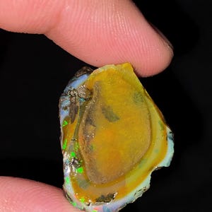 以下が含まれることがあります： 鮮やかな黄色とオレンジ色の中央部が特徴の、未研磨の原石オパール。半透明の乳白色の縁取りに囲まれており、端には暗い岩石の母岩を背景に、明るい緑色と青色の光の虹色の閃光が見られます。