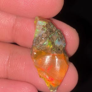 以下が含まれることがあります： 鮮やかなオレンジと赤のベースカラーに、緑と黄色の虹色の輝きを放つ、未研磨の原石オパール。石は粗く不規則な形状をしており、表面には暗い土のような母岩の斑点が埋め込まれています。