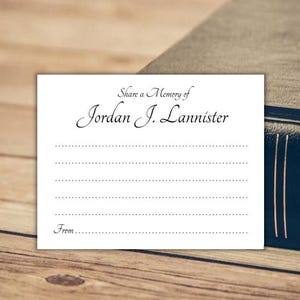 May include: A white card with the text "Share a Memory of Jordan J. Lannister" in elegant script. Below the text are dotted lines for writing, with "From..." at the bottom. The card rests on a wooden surface next to a dark blue book.