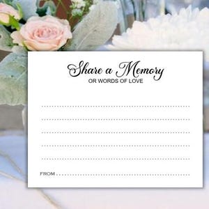 May include: A white card with the text "Share a Memory OR WORDS OF LOVE" in elegant script. The card has dotted lines for writing and a space for "FROM...". Soft pink roses and greenery are in the background.