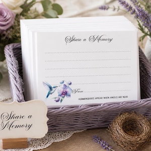 May include: A stack of white rectangular memory cards featuring the text Share a Memory, a hummingbird and orchid illustration, and the phrase HUMMINGBIRDS APPEAR WHEN ANGELS ARE NEAR, placed inside a light purple woven basket with a matching sign.