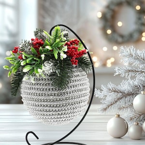 Puede incluir: Una maceta decorativa plateada en forma de esfera con un soporte de metal negro, llena de vegetación artificial y bayas rojas. La maceta tiene un diseño texturizado y tejido. Adornos navideños blancos y una rama helada en el fondo.
