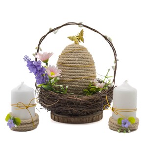 Puede incluir: Centro de mesa decorativo con forma de colmena tejida con una mariposa dorada en la parte superior, anidada en un nido de ramitas. Dos velas de pilar blancas con detalles florales flanquean el centro de mesa, creando una est&eacute;tica r&uacute;stica.