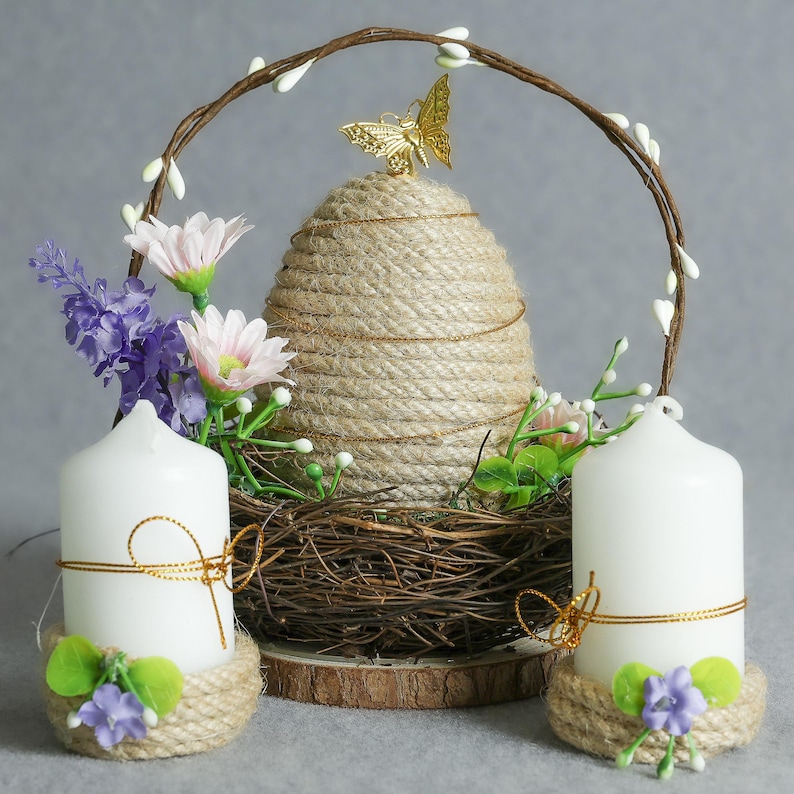 Puede incluir: Centro de mesa decorativo con forma de colmena tejida, coronado por una mariposa dorada, flanqueado por velas blancas, flores y un arco de ramitas. Las velas tienen detalles de cuerda y flores, creando una exhibici&oacute;n r&uacute;stica y elegante.