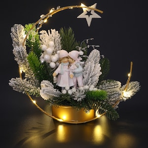 May include: A decorative Christmas centerpiece featuring a gold-colored crescent-shaped frame with a star and fairy lights. The centerpiece includes a miniature figurine of two children, artificial snow-covered pine branches, and white berries.