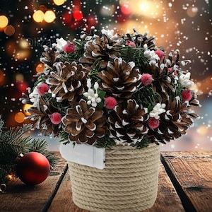 May include: A Christmas decoration comprising a sphere of brown pine cones, red berries, and white accents, set in a rope-wrapped pot. A red bauble and evergreen branch are in the foreground, with a blurred background of lights and falling snow.