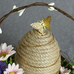 Puede incluir: Centro de mesa decorativo con base de cuerda enrollada, adornado con una mariposa dorada y una cadena dorada. El arreglo incluye flores artificiales en tonos rosa, morado y blanco, con un arco de ramitas marrones.