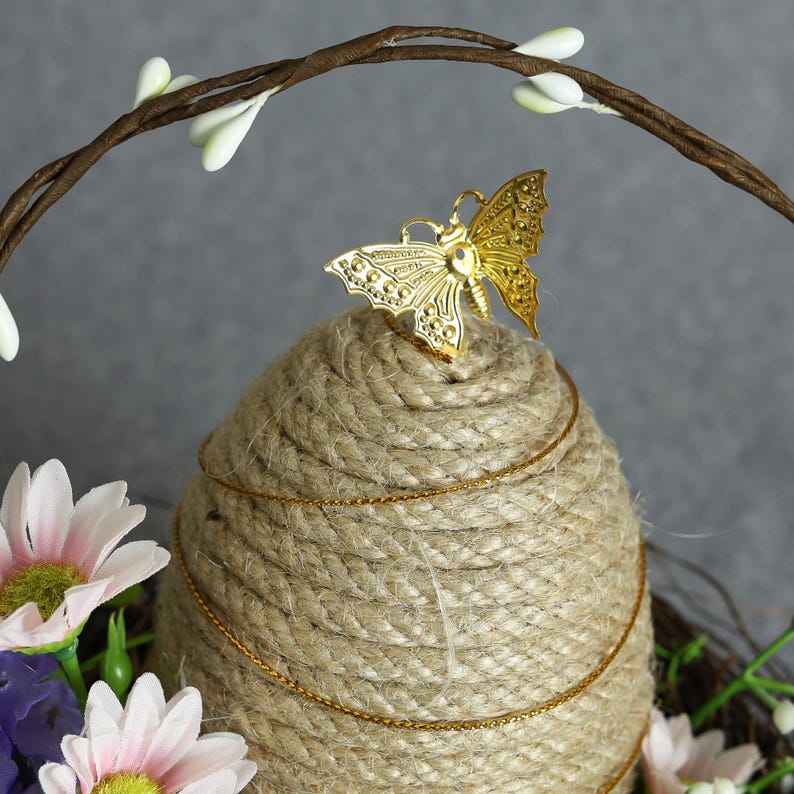 Puede incluir: Centro de mesa decorativo con base de cuerda enrollada, adornado con una mariposa dorada y una cadena dorada. El arreglo incluye flores artificiales en tonos rosa, morado y blanco, con un arco de ramitas marrones.