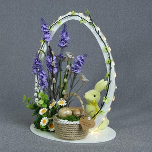 May include: An Easter decoration with a white arch decorated with lights and foliage. A yellow bunny sits next to a basket filled with eggs and flowers. The basket has a heart-shaped tag that says "Handmade".