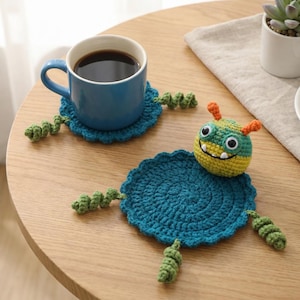 Könnte beinhalten: Eine blaue Keramiktasse mit Kaffee steht auf einem passenden blauen gehäkelten Untersetzer mit grünen Ranken. Ein zweiter Untersetzer mit einem gehäkelten Monstergesicht ist ebenfalls vorhanden. Die Untersetzer stehen auf einem hellen Holztisch.