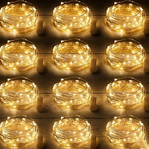 May include: A collection of coiled, warm white LED string lights. Each coil is paired with a small, cylindrical battery pack. The lights are arranged on a light-colored wooden surface, creating a cozy, ambient glow.