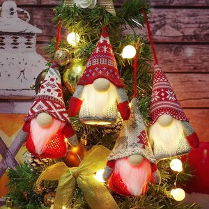 May include: Four Christmas gnome ornaments with red and white knit hats and glowing white beards. The gnomes have red mittens and gray sleeves. They are hanging on a Christmas tree with lights and a gold bow.