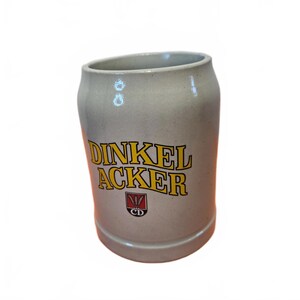May include: A light grey ceramic beer mug with a smooth, cylindrical body. The front features the text DINKEL ACKER in bold yellow letters with black outlines, positioned above a small red shield logo containing three wheat stalks and the letters CD.