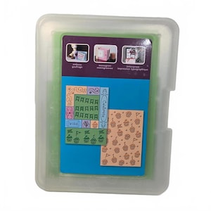May include: A translucent plastic storage case containing craft supplies. The insert features a purple header with text including "emboss", "monogram", and "letterpress". Below are two rectangular panels showing printed designs of cupcakes, banners, and the word "Celebrate".