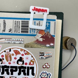 May include: A white bookmark with the word "Japan" in red, attached to a journal. The journal features stickers with Japanese themes, such as Mount Fuji, trains, and geishas. The bookmark is made of plastic.