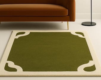 Puede incluir: Una alfombra rectangular verde con un borde crema y recortes decorativos en las esquinas. La alfombra está colocada sobre un suelo blanco, con un sofá marrón, una lámpara de pie negra y una pintura azul oscuro en el fondo.