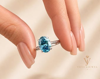 May include: A silver ring featuring a large, oval-shaped, light blue gemstone. The ring is held between two fingers against a soft, neutral background. The ring's band is thin, emphasising the gemstone's colour and shape.