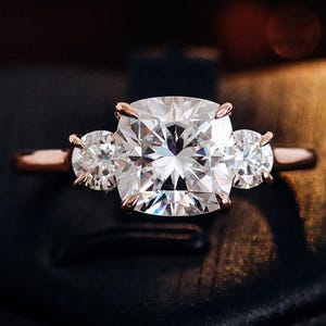 Può includere: Un anello in oro rosa con una grande pietra centrale a taglio quadrato affiancata da due pietre rotonde più piccole. L'anello è incastonato su uno sfondo scuro, evidenziando la brillantezza delle gemme chiare. Un classico anello di fidanzamento a tre pietre.