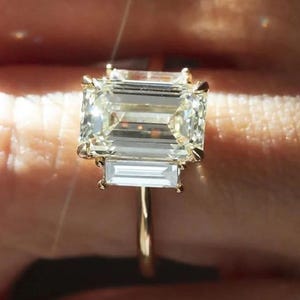 May include: A gold ring with a large emerald-cut diamond as the central stone, accompanied by two smaller, rectangular diamonds. The ring has a slender, polished gold band. The design accentuates the sparkle of the clear, brilliant diamonds.