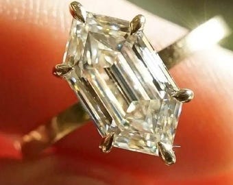 May include: A gold ring featuring a large, clear, geometric-cut diamond. The diamond has a rectangular shape with angled sides and is held in place by small prongs. The ring is held by a person's fingers.