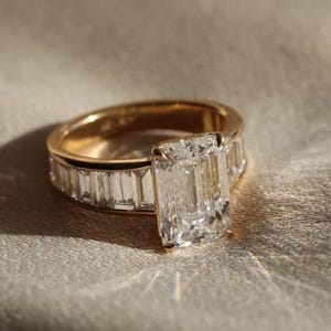 May include: A gold ring with a large, rectangular-cut diamond center stone. The band is also adorned with smaller, rectangular-cut diamonds. The ring is set against a neutral, textured background, highlighting its sparkle.