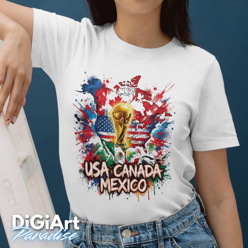May include: White t-shirt featuring a colorful graphic design. The design includes a gold trophy, the flags of the USA, Canada, and Mexico, and the text "USA CANADA MEXICO". The design is set against a backdrop of paint splatters.